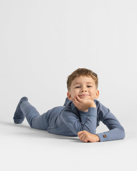 Boy lying prone on the floor propping his chin up with his hands, wearing a complete dusty blue thermal set.