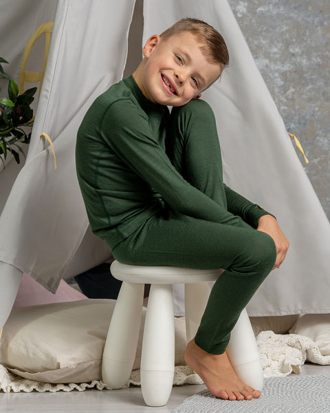 Child in a dark green merino wool outfit sitting on a white stool in a cozy indoor setting with a teepee.