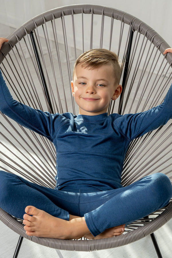 A boy wearing the menique Kids' 160 Merino Pants in Denim color. He is sitting in a lounge chair. 