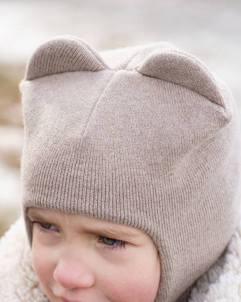 A close-up of a child&#39;s head, partially obscured, wearing a creamy beige menique knit balaclava or hat with two ear-like protrusions on top. The child&#39;s right eye and part of their face are visible, and they appear to be looking slightly to their right. A textured, light-colored collar or hood of a jacket is visible around their neck, and the background is blurred with light tones, suggesting an outdoor setting.