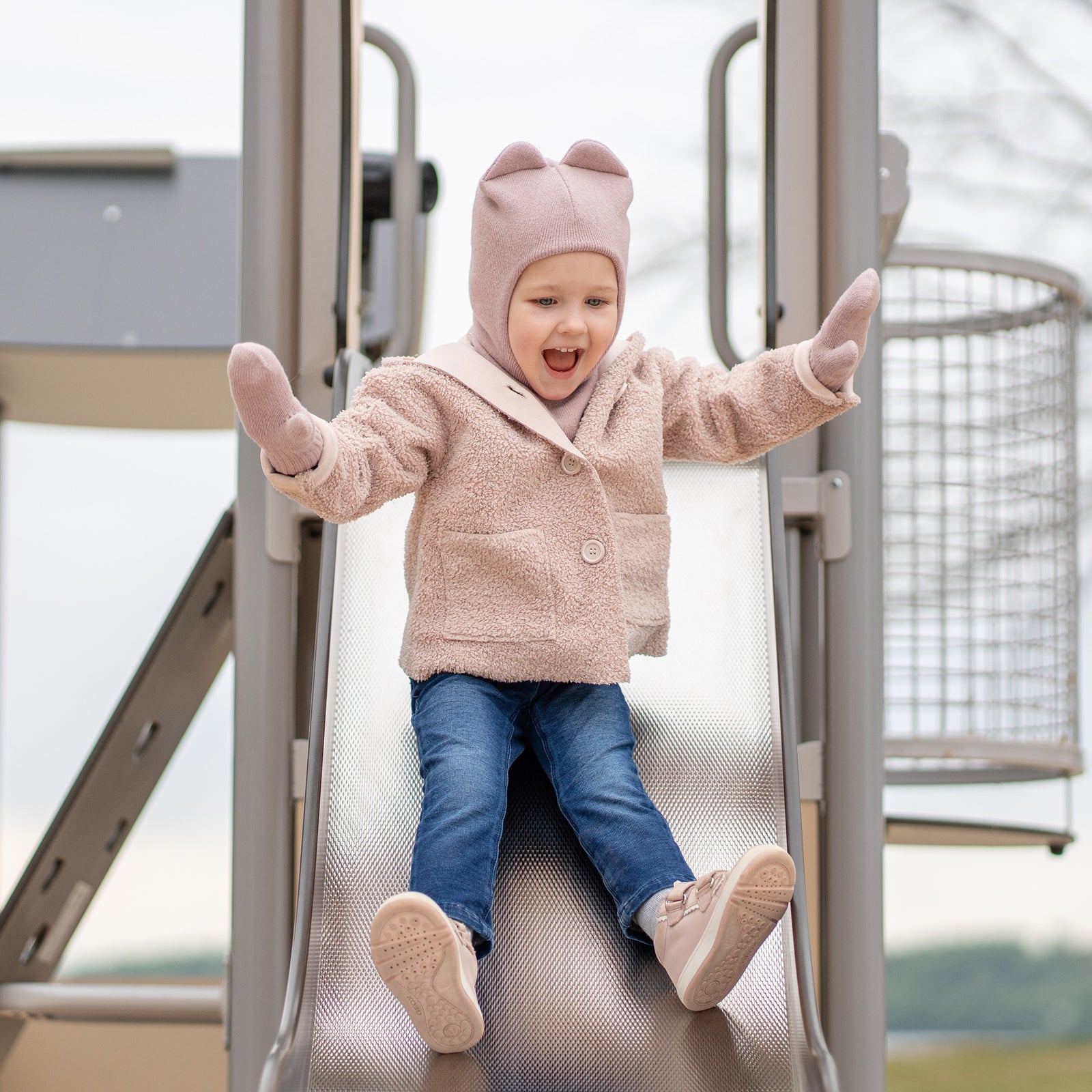 A young child wearing a light blue balaclava with small ears, light blue mittens, and a grey and black outfit, smiles while looking at the viewer. They are partially obscured by a metal swing chain on the left, with a blurred sandy ground in the background.
