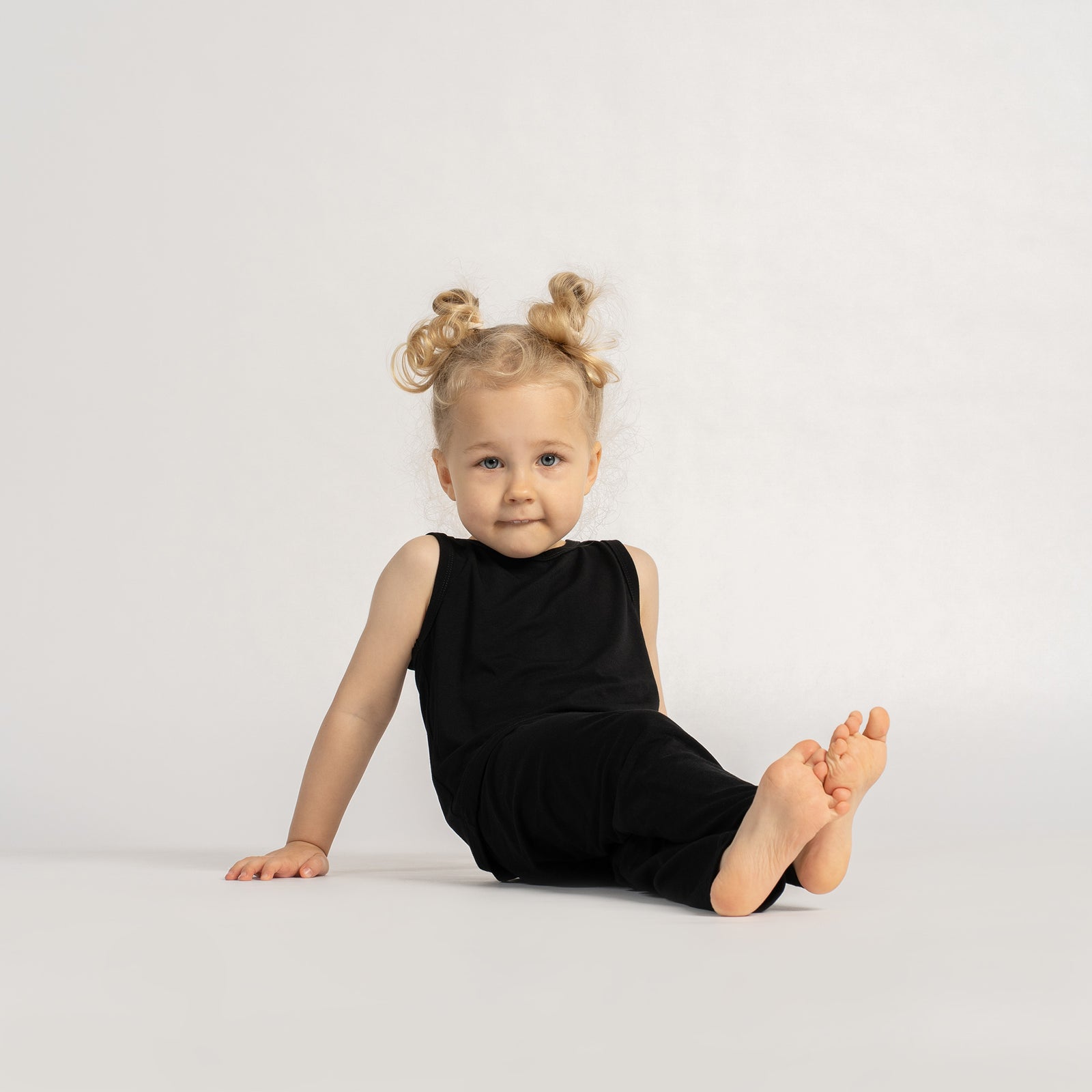 Smiling little girl in a black sleeveless top and leggings, standing barefoot with hands behind her back against a neutral background.