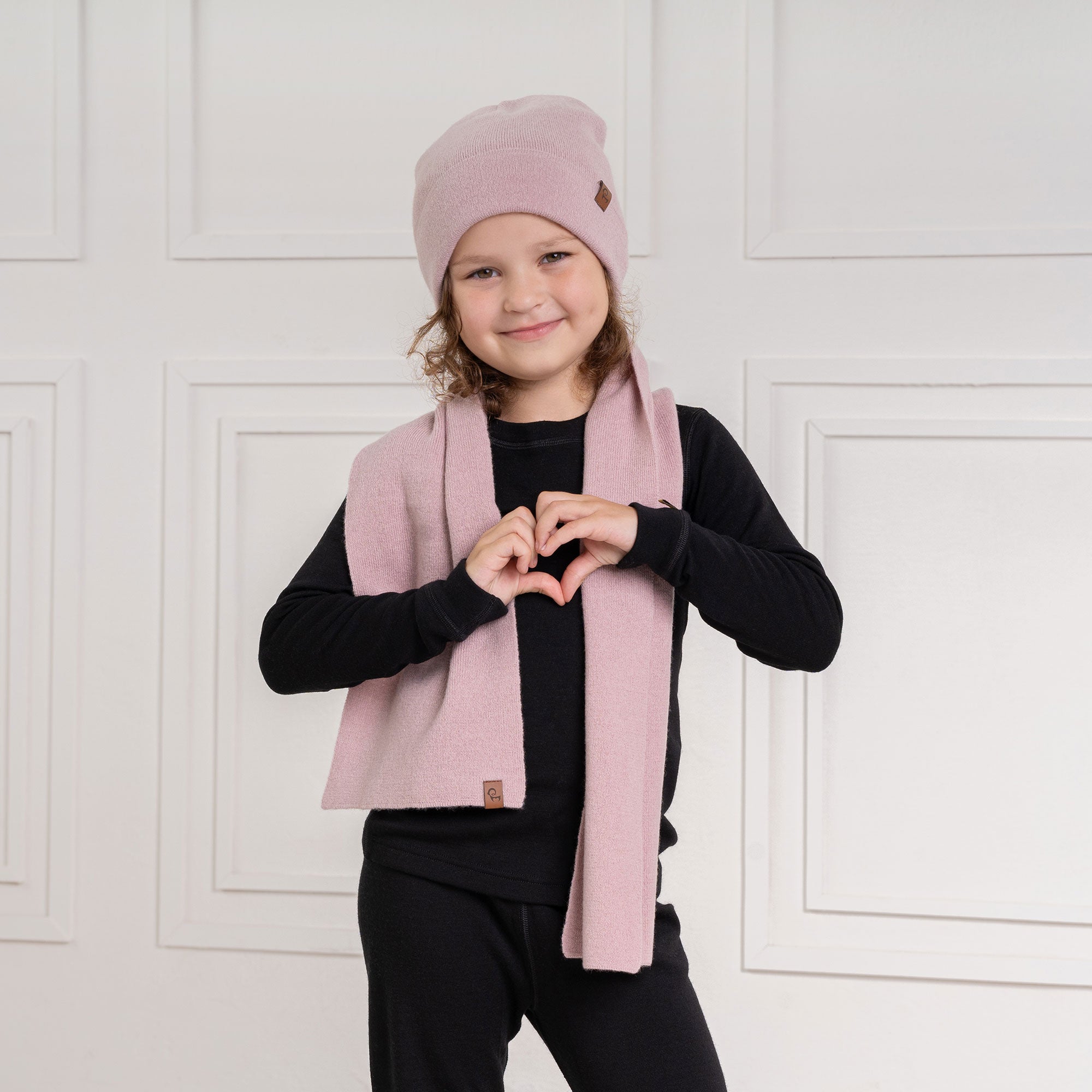 A young child with curly brown hair and a slight smile looks directly at the viewer. They are wearing a black long-sleeved shirt and a light pink scarf with a small brown label, draped around their neck. The background is a white paneled wall.