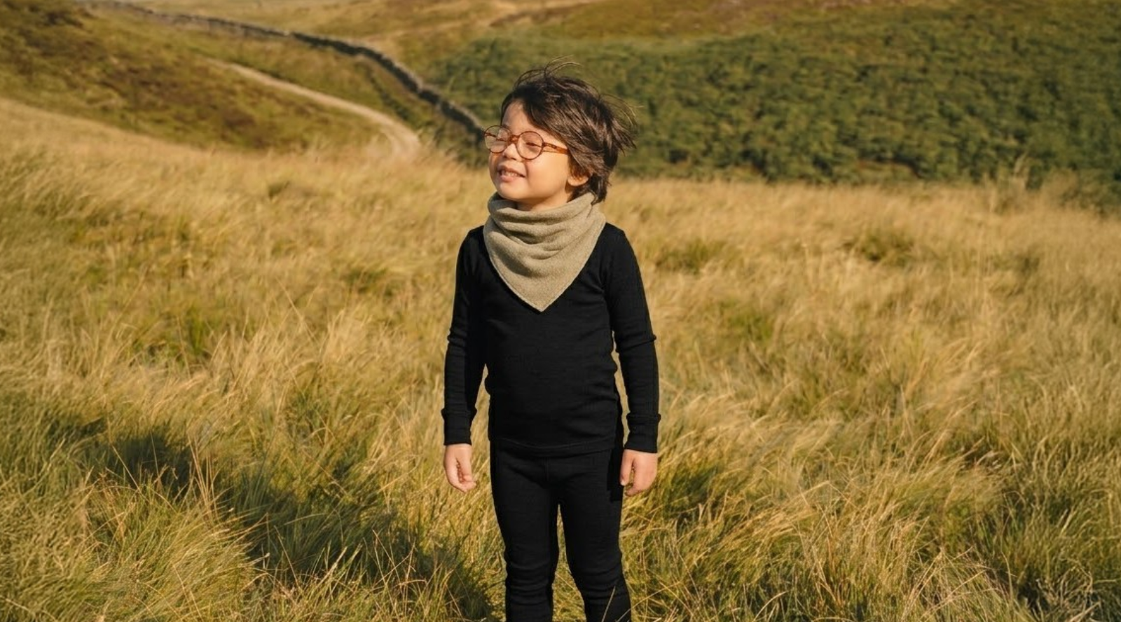 A young child wearing glasses, a black long-sleeved top and pants, and a beige neck warmer, standing in a golden grassy field with rolling hills and a dirt path in the background.