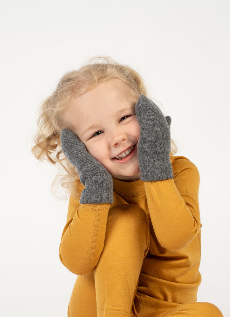 A cheerful young child with blonde curly hair smiling brightly, wearing a mustard yellow long-sleeved top and framing their face with hands clad in grey knit mittens.