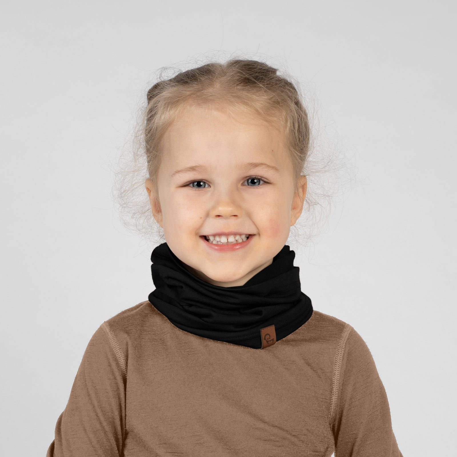 A studio portrait of a smiling young blonde girl looking off to the left side of the frame. Her blonde hair is pulled back into a ponytail. She is wearing a dusty pink neck gaiter gathered around her neck, which has a small brown leather logo patch near the bottom edge. She is wearing a solid black long-sleeved shirt against a plain light grey background.