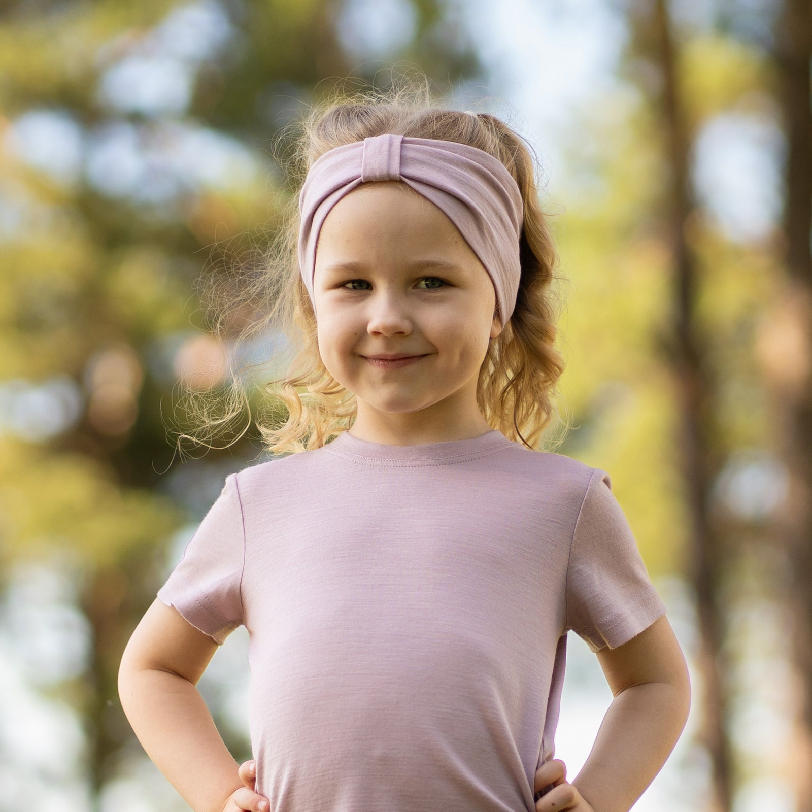 Young girl smiling outdoors wearing a dusty pink Merino wool T-shirt and a matching Merino wool headband. The soft, breathable accessories and clothing are showcased in a sunny natural setting, offering comfortable everyday wear for active ki