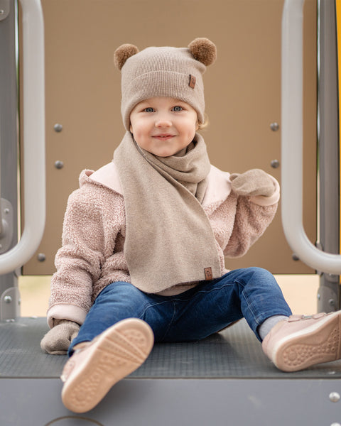 A young child with a sweet smile, sits on a playground structure. They are wearing a beige balaclava with brown pom-pom ears, a matching beige scarf, a light pink sherpa-style coat, blue jeans, and pink sneakers. Their eyes are looking towards the camera.