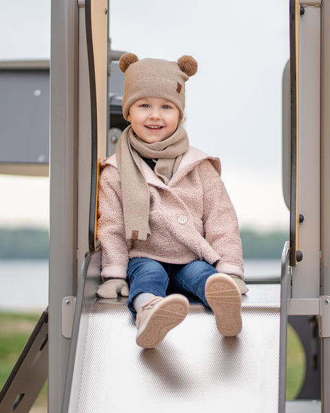A young child with a cheerful smile, is sitting at the top of a metal playground slide. They are dressed in a beige balaclava with brown pom-pom ears, a beige scarf, a light pink sherpa-style coat, blue jeans, and light pink sneakers. A blurred background shows water and trees.