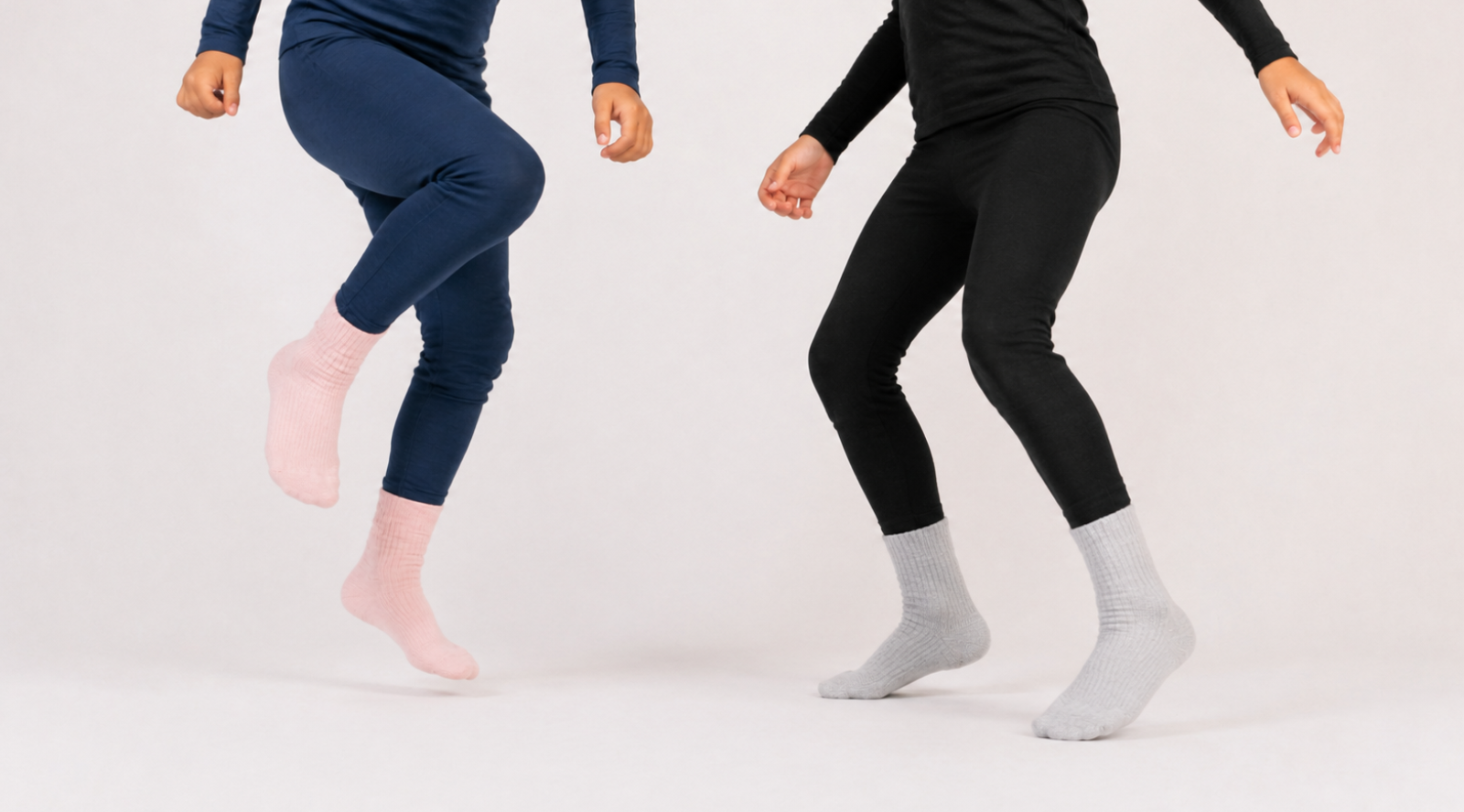 The lower halves of two people jumping in front of a white backdrop. The individual on the left wears navy blue leggings with pink ribbed socks, and the individual on the right wears black leggings with grey ribbed socks.