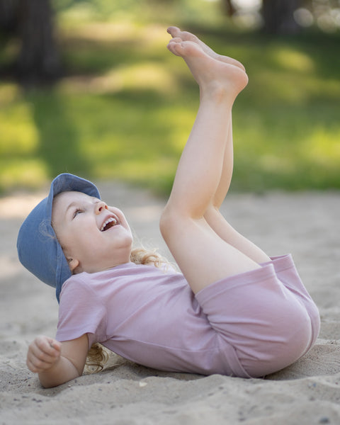 Young girl sitting on a sandy beach wearing a dusty pink Merino wool T-shirt, matching shorts, and a dusty blue sun cap hat. The lightweight, breathable kids' outfit is showcased in a sunny outdoor setting, making it ideal for summer adventures, travel, and everyday comfort.