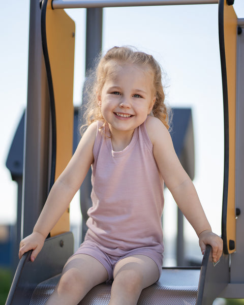A cheerful young girl sitting at the top of a playground slide, holding onto the sides and facing forward with a bright smile. She has blonde hair and is wearing a dusty pink tank top and shorts set.