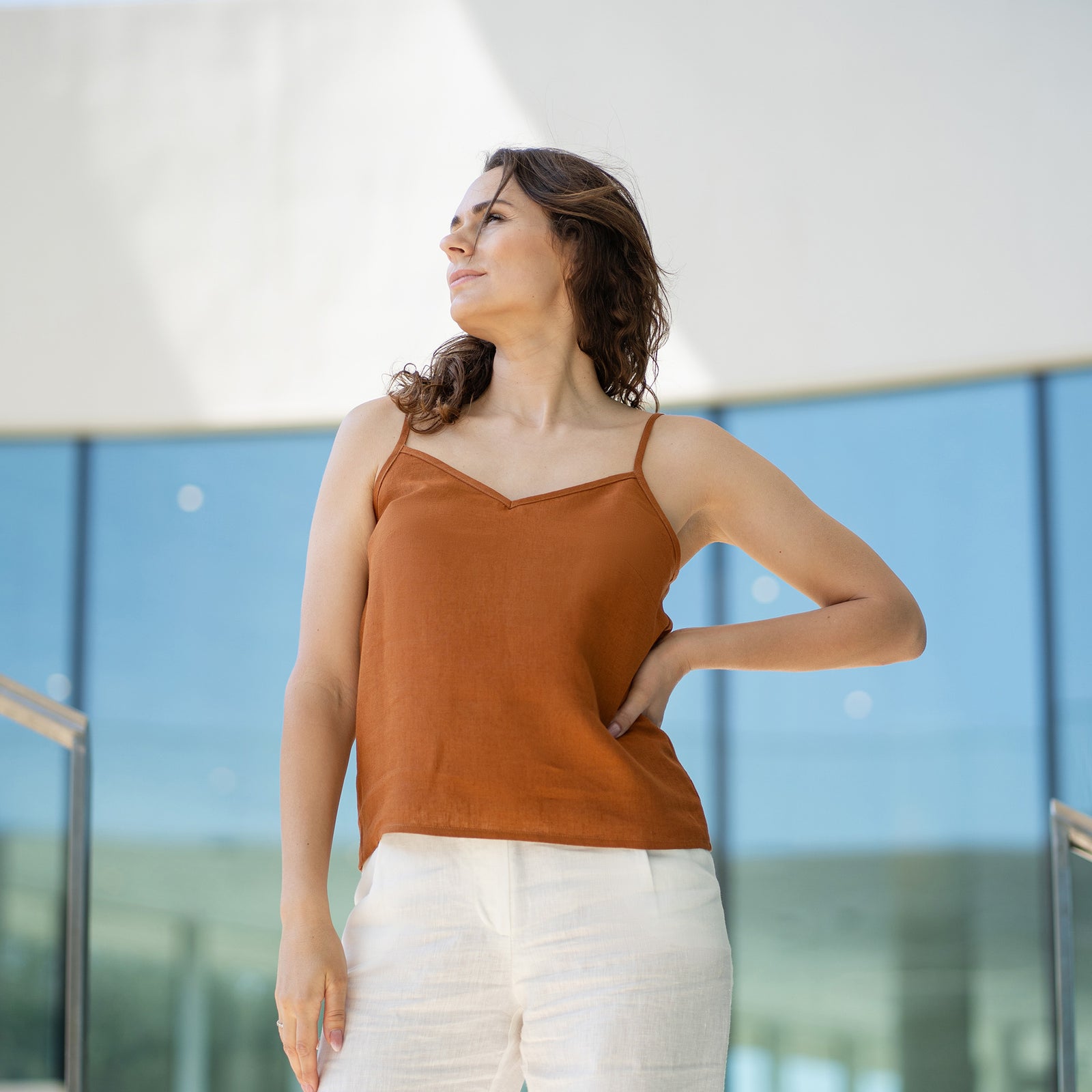A woman with brown hair wears a almond brown V-neck camisole top and white trousers, posing indoors in front of large glass windows.