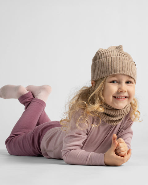 A young girl lying on her stomach propped on her elbows, wearing a dusty rose two-piece loungewear set and a light beige knit beanie.
