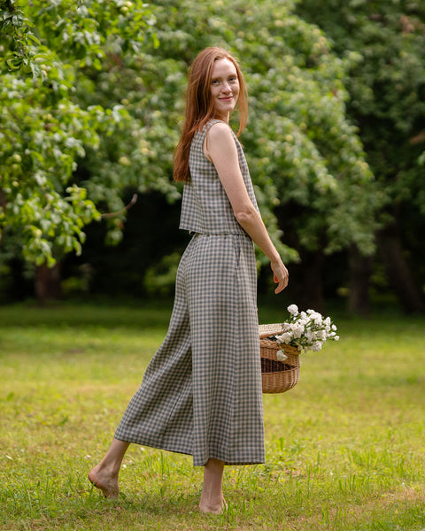 The model is standing sideways, with her back also showing. She is wearing the Lana green gingham culottes matching with a crop top in the same color pattern. She is smiling lightly, holding a flower basket. One of her legs is moved slightly backward, so the back of the LANA culottes is visible.