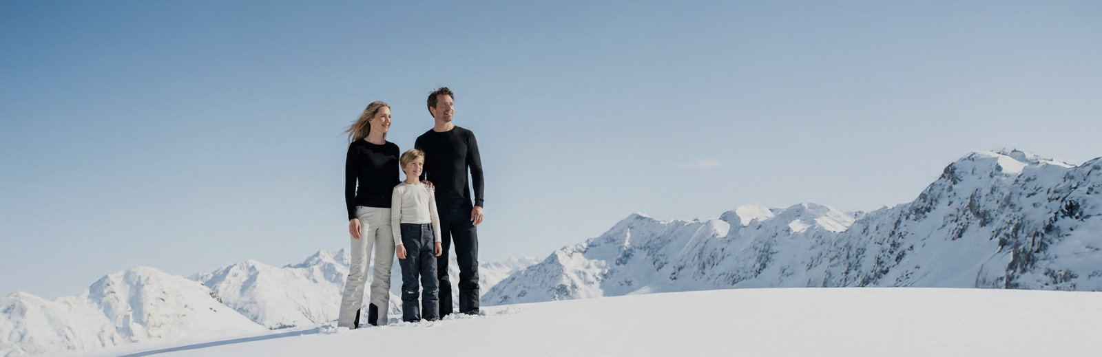 Parents and children smiling on a snow-covered mountain, dressed in matching black base layers and insulated pants under a clear blue sky.