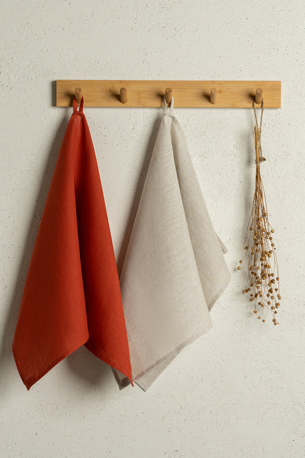 Two menique linen kitchen dish towels hanging on a wooden rack on a wall. The colors of the towels are cinnamon red and natural. 