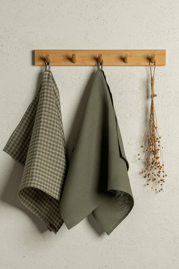 Two menique linen kitchen dish towels hanging on a wooden rack on a wall. The colors of the towels are stone green and green check. 