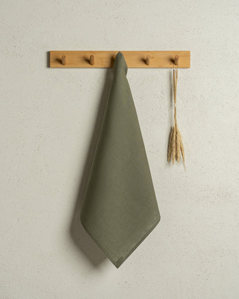 The menique square linen dish towel in color stone green hanging on a wooden rack on a wall.