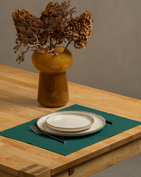 The menique linen placemat in dark green color on a wooden table top. There is a vase on top of the table, as well as some cutlery by the sides of the plate, that is on top of the placemat.