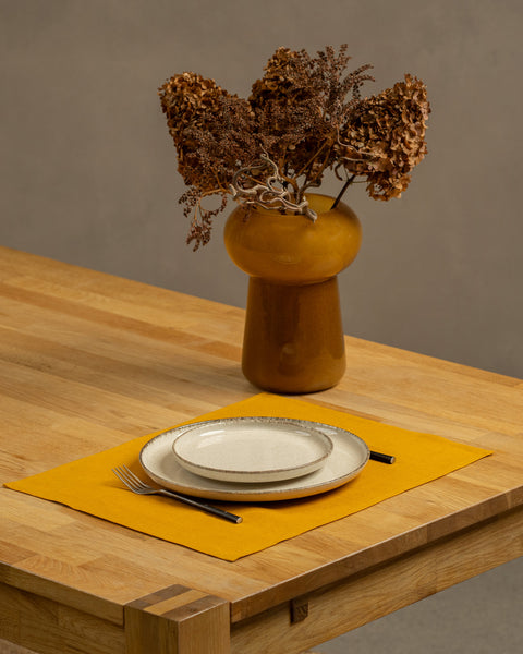 The menique spicy yellow linen placemat on a light wooden table. There are two different size porcelain plates and cutlery on top of it. There is also a vase on the table.