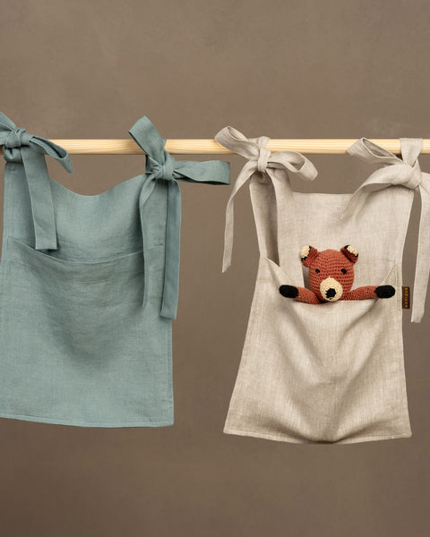 Two linen baby bed organizers hanging side-by-side from a light wooden bar. The organizer on the left is a dusty sage green, and the one on the right is a natural light beige. Both feature wide fabric ties secured with bows over the bar and a large front pocket. The beige organizer has a small, crocheted brown bear toy peeking out of its pocket. The background is a solid, warm taupe color.