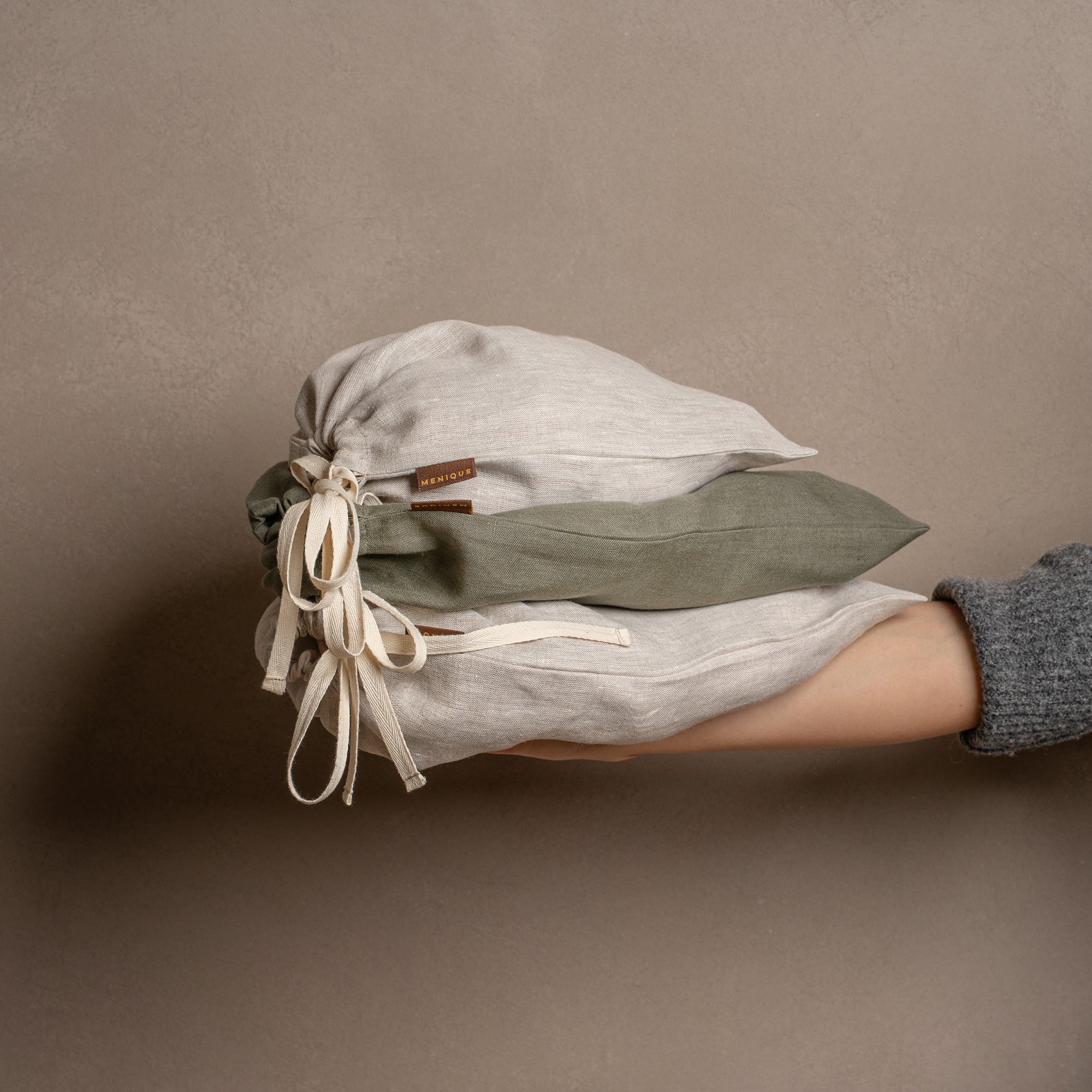 Five closed linen drawstring bags in varying sizes and colors of natural and stone green are arranged on a textured jute rug against a warm brown wall.
