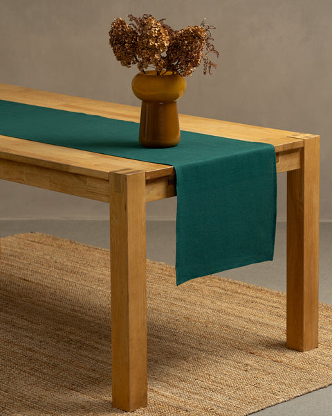 A wooden table positioned on top of a carpet. There is a dark green linen table runner and a vase on top.