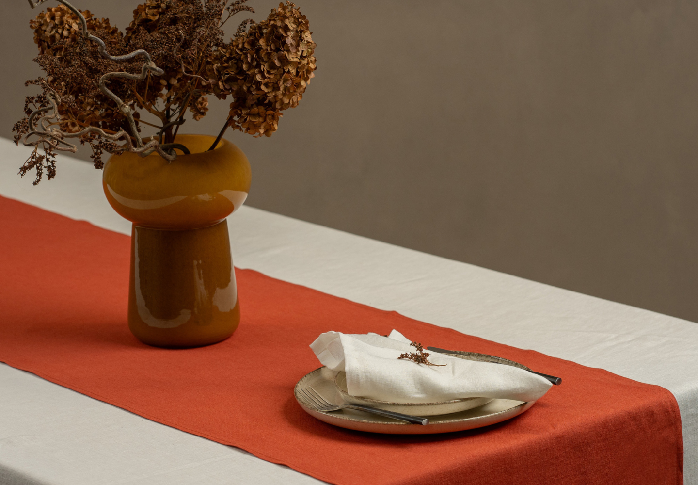 A minimalist table setting featuring a cinnamon red linen table runner from menique on a cream-colored tablecloth, with a ceramic vase of dried flowers and a neutral-toned plate set with a folded linen napkin and dried floral accent.