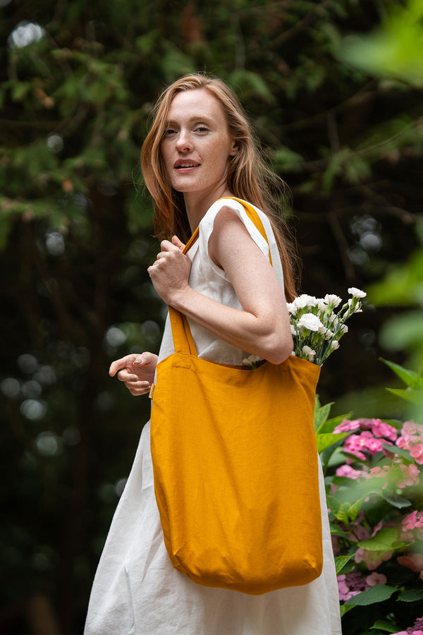 A fair-skinned woman with long, reddish-blonde hair looks over her shoulder with a gentle smile. She is wearing a light-colored dress and carrying a spicy yellow tote bag filled with white flowers. The background features blurred green foliage and pink flowers, suggesting an outdoor setting.