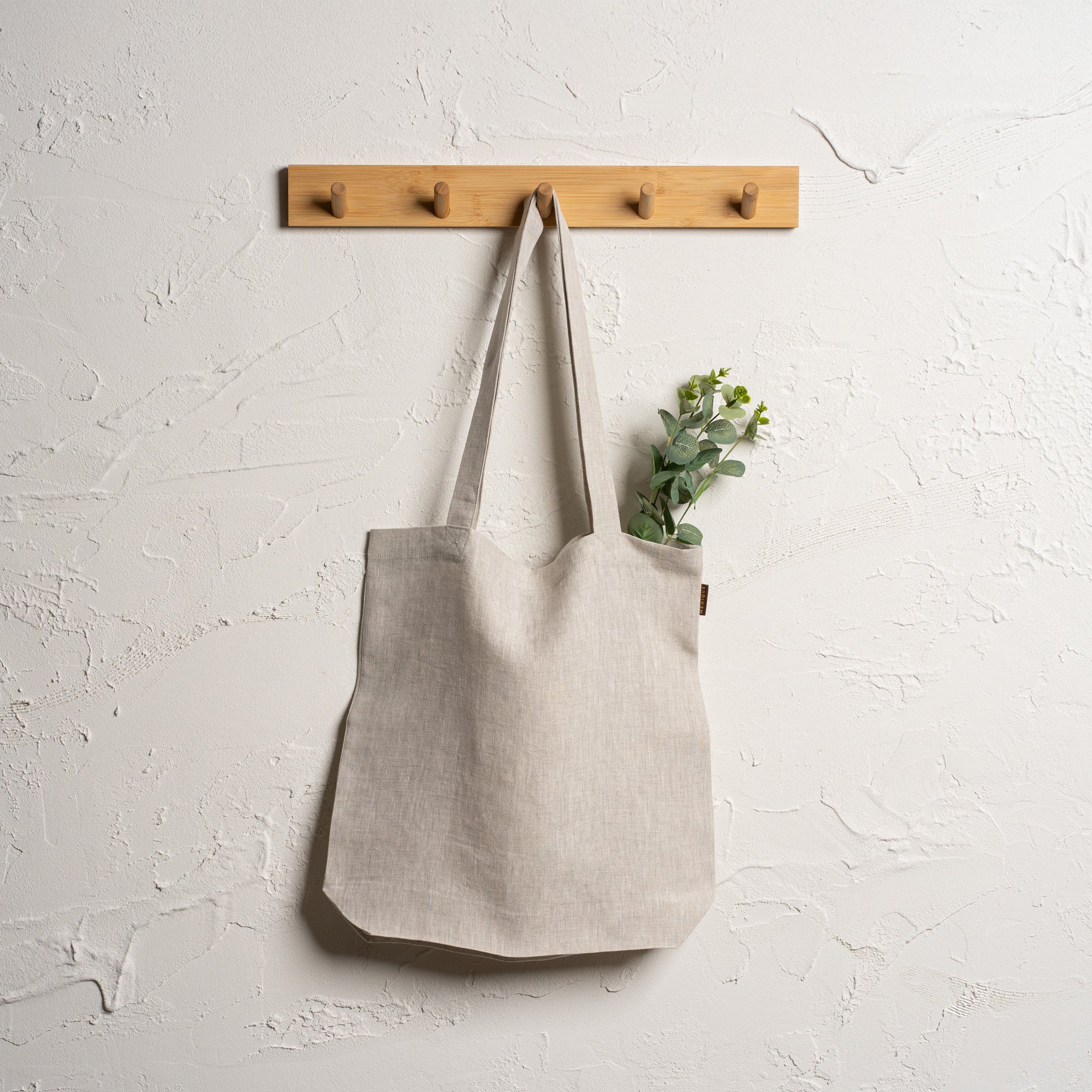 Menique natural color linen tote bag with long handles hangs from a branch of a light-colored tree. In the blurred background, there is a body of water reflecting the sky, surrounded by green grass and trees under a partly cloudy sky.
