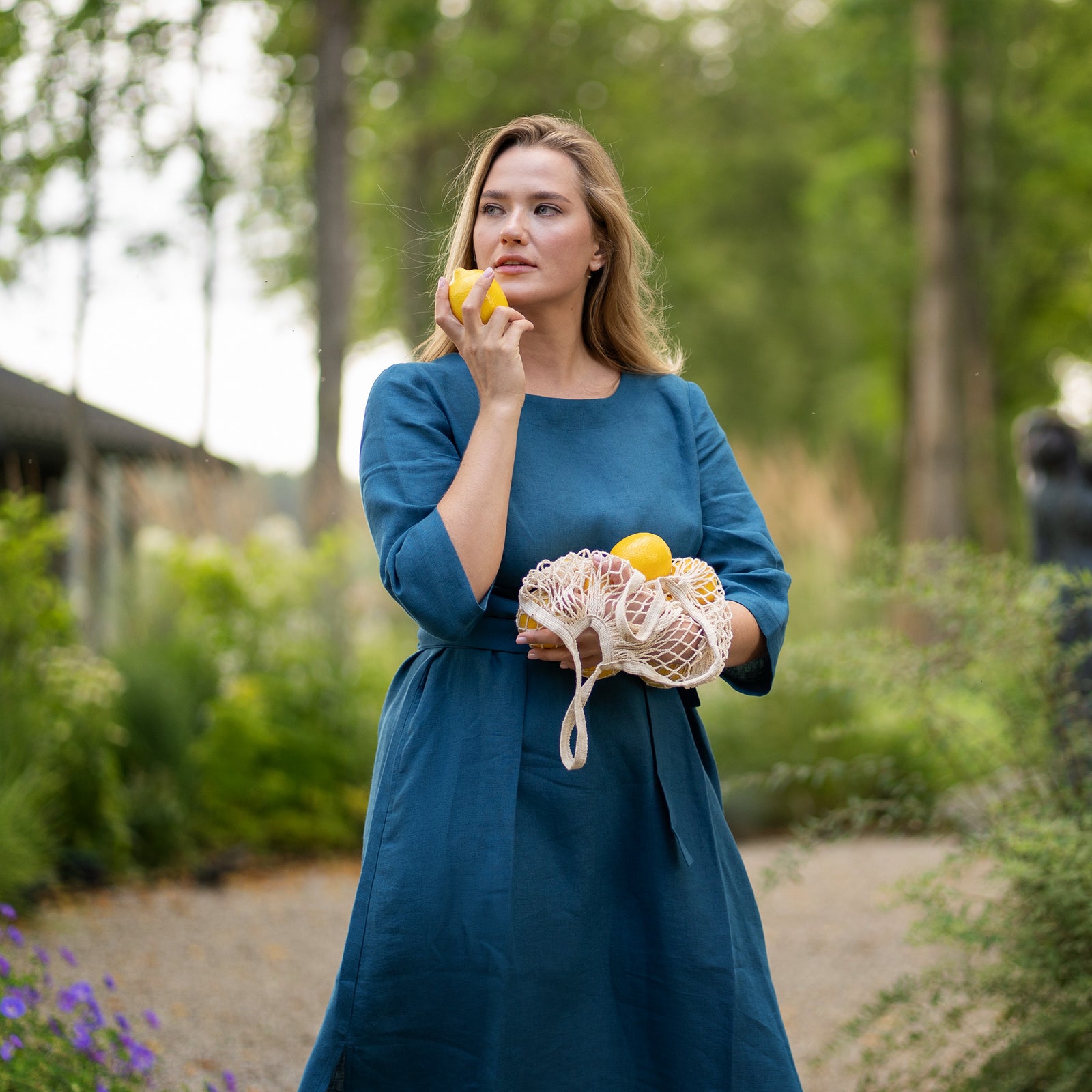 A smiling woman stands on a gravel garden path wearing a cobalt blue, three-quarter sleeve linen dress with a tied waist. She playfully holds up a single lemon in one hand while carrying a string bag full of lemons in the other.