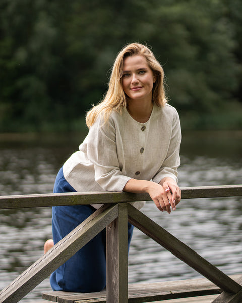 A woman leans comfortably against a wooden pier railing with a calm, tree-lined lake in the background. She is wearing a natural-colored, button-up linen jacket and storm blue linen pants, smiling softly at the viewer.
