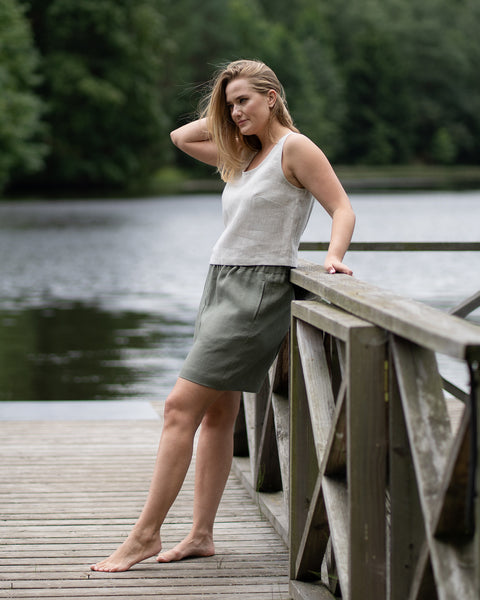 A barefoot woman stands on a wooden pier, casually leaning against the railing while resting one hand in her hair. She is dressed in a natural linen tank top and stone green bermuda shorts, with a tranquil lake and forest setting the scene behind her.