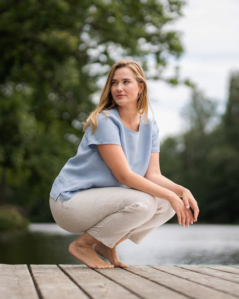 A woman in a light blue short-sleeved blouse and light beige pants crouches on a wooden pier. She rests her arms on her knees while looking off to the side against a serene backdrop of green trees and water.