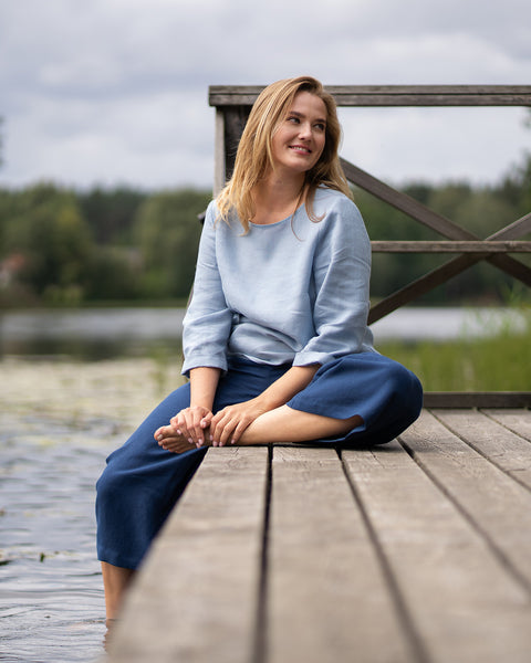 A woman sits cross-legged on a wooden pier next to a lake, resting her hands on her bare foot while smiling warmly toward the distance. She is dressed comfortably in a cloudy blue long-sleeve linen top and relaxed, storm blue linen pants.