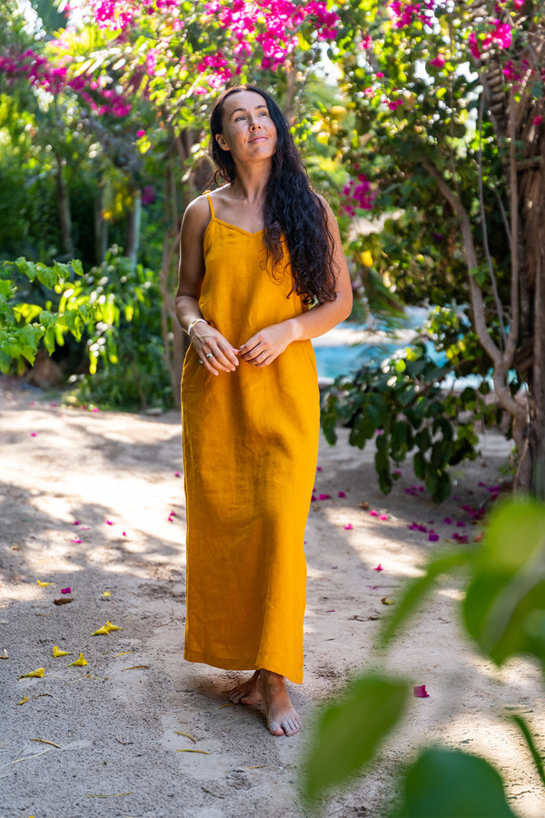 A woman with long, dark, wavy hair stands barefoot on a sandy path, looking up towards the left. She is wearing a mustard yellow linen sundress with thin straps. Lush green plants and trees surround her, and there are pink flowers blooming in the background. Yellow petals are scattered on the path.