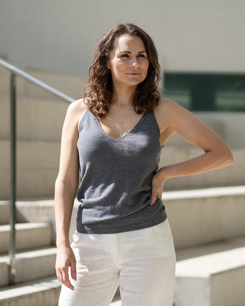 A woman with wavy brown hair looks to her right, hand on hip, wearing a dark gray tank top and white menique trousers. She stands on light-colored stairs with silver railings.