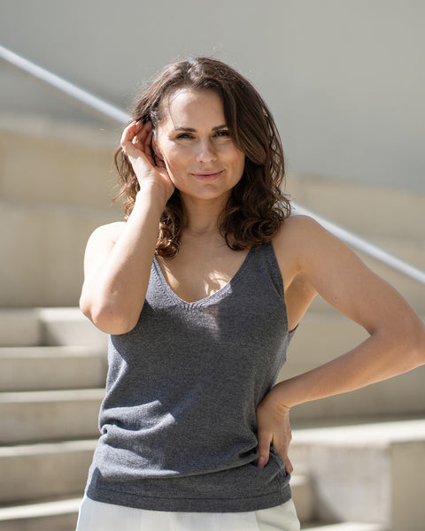 A woman with shoulder-length wavy brown hair smiles gently at the camera, with one hand near her ear and the other on her hip. She is wearing a dark gray v-neck tank top and pure white trousers, standing on light-colored steps with a silver handrail, in what appears to be natural daylight.