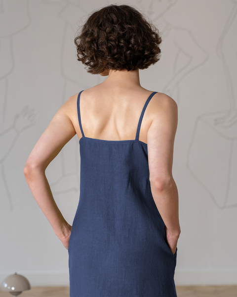 A shot from behind shows a woman with short, curly dark hair, wearing a sleeveless, square-neck, storm blue dress with thin straps and pockets. Her hands are in the pockets. A light-colored wall with faint white line art is in the background, and a small, gray dome-shaped lamp is partially visible in the lower left corner.