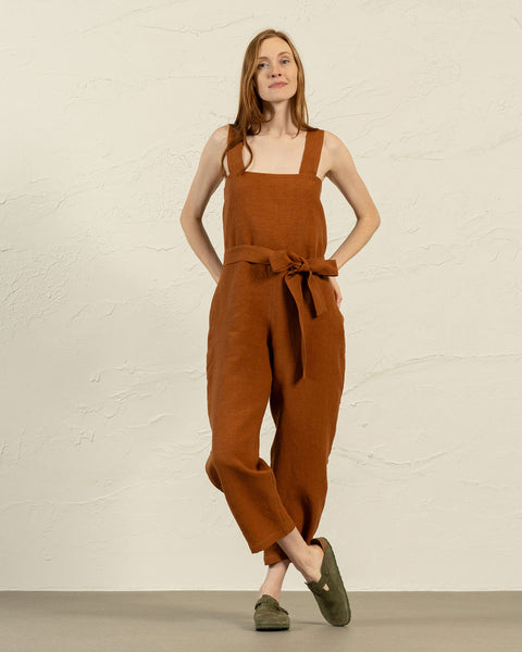A light-skinned woman with long brown hair stands facing forward, wearing a linen menique jumpsuit with a tied belt at the waist and olive-green slip-on shoes. She has a neutral expression and her hands are resting on her hips.