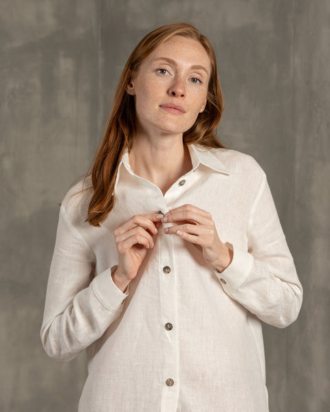 Woman with reddish hair wears a buttoning white linen shirt against a gray background