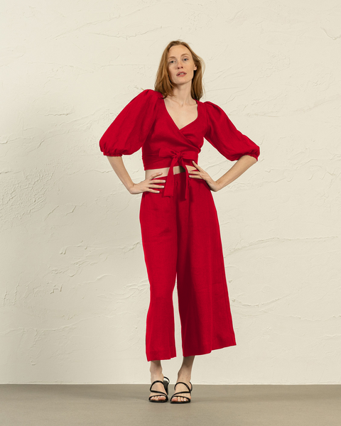 A fair-skinned woman models a pure red two-piece outfit against a textured off-white wall. She stands facing forward with her hands resting on her hips, exuding confidence. The outfit consists of menique wrap top with a deep V-neck and voluminous three-quarter sleeves gathered at the elbows. The top ties at the waist, accentuating her figure and revealing a glimpse of her midriff.