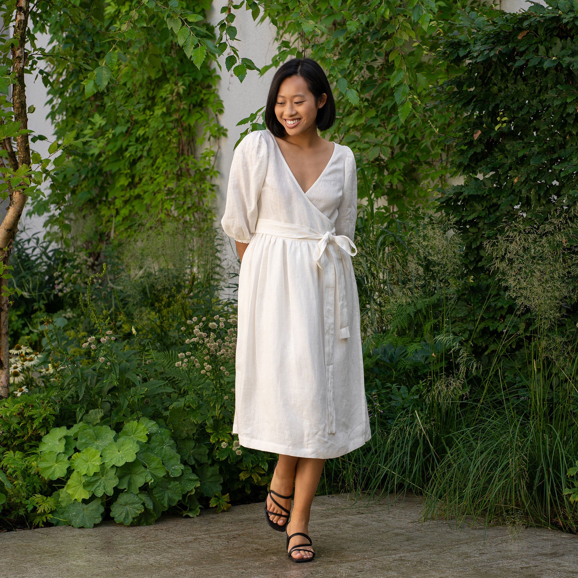 A smiling person with short, dark hair and what appears to be Asian features is holding a bouquet of light pink and white flowers. They are wearing a pure white wrap menique dress with elbow-length sleeves and are standing outdoors amidst lush green foliage and other flowers.