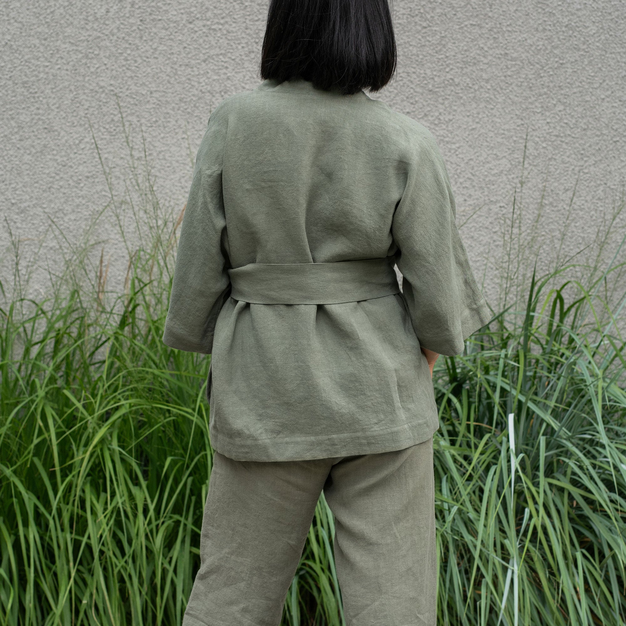A person stands outdoors, wearing a stone green colored linen kimono-style top and matching wide-leg pants. Their right hand is in the pocket of their pants. To their left, there are tall green plants.