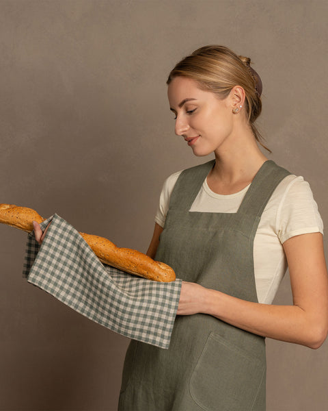 A woman with blonde hair holds a baguette wrapped in a menique linen cloth, wearing a stone green linen cross-back apron, against a neutral background.