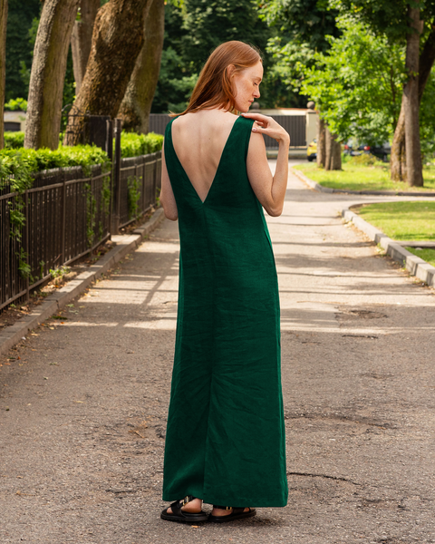 A woman with long reddish hair, wearing a menique sleeveless dark green linen dress with a low V-back, stands on a paved path in a park with trees and greenery on either side. She is looking down and to the left, with her right hand gently touching her left shoulder.