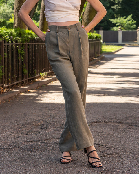 A person is shown from the waist down, modeling stone green, high-waisted, linen wide-leg trousers. They are wearing black open-toe sandals with thin straps and have their hands on their hips. The setting appears to be outdoors on an asphalt path, with a black metal fence and greenery visible in the background.