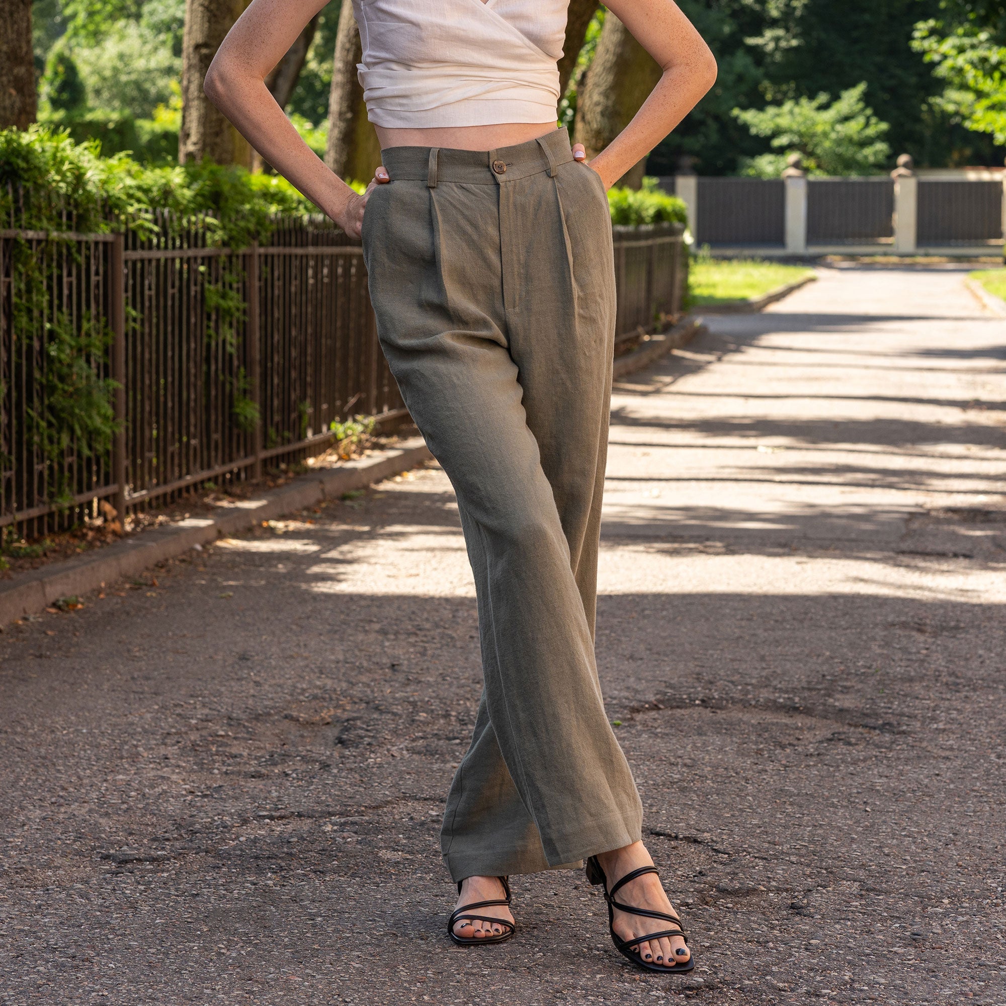 A fair-skinned woman models a cinnamon red colored linen vest and wide-leg trousers, standing against a textured light beige wall. She is wearing black sandals.