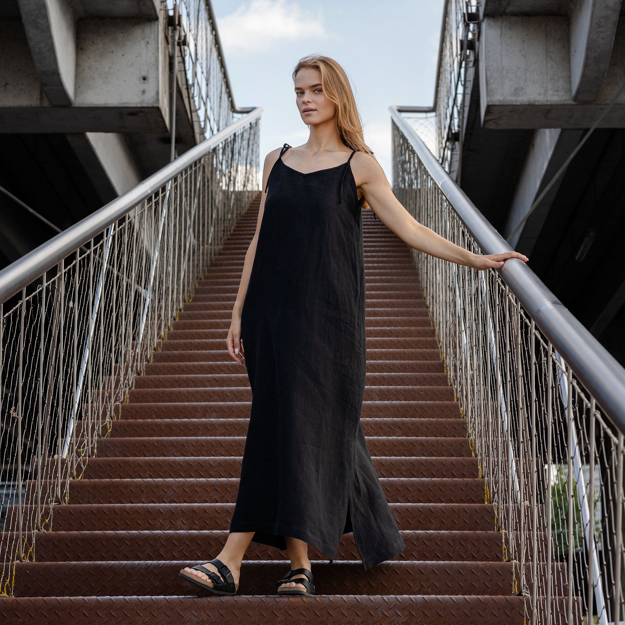 A light-skinned person with long blonde hair is standing on a set of outdoor stairs, looking towards the camera with a gentle smile. They are wearing a pure black sleeveless linen dress with thin straps, and one hand is resting on their chest while the other holds onto a metal railing. The background shows a cloudy sky and architectural elements of a walkway or bridge.